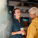 Two women colleagues laughing while standing in a cafe at their workplace. One of the women is holding a take out hot drink cup.