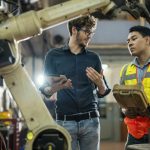 Software engineer explaining to controlling robotic welding process to welder in factory.
